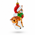 A glass Christmas ornament shaped like a brown deer wearing a red Santa hat with white trim and a green bow tie with a red bow, shown on a white background.