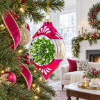 A festive Christmas ornament hanging on a decorated tree in a cozy living room. The ornament is a glossy magenta pink teardrop shape with white cutout leaf patterns, a clear lower half, and a central green glittery floral design.