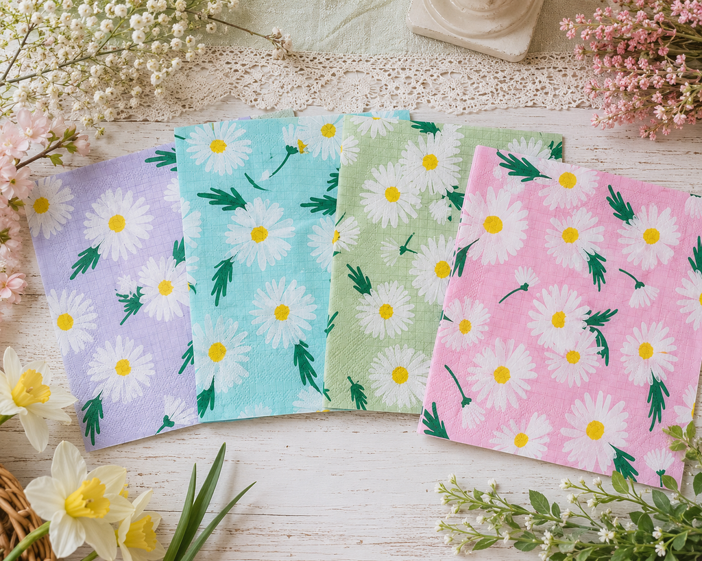 Four square paper napkins with a daisy pattern are arranged in a fan shape on a white wooden surface. The napkins come in four colors: lavender, light blue, light green, and pink. Great for crafts