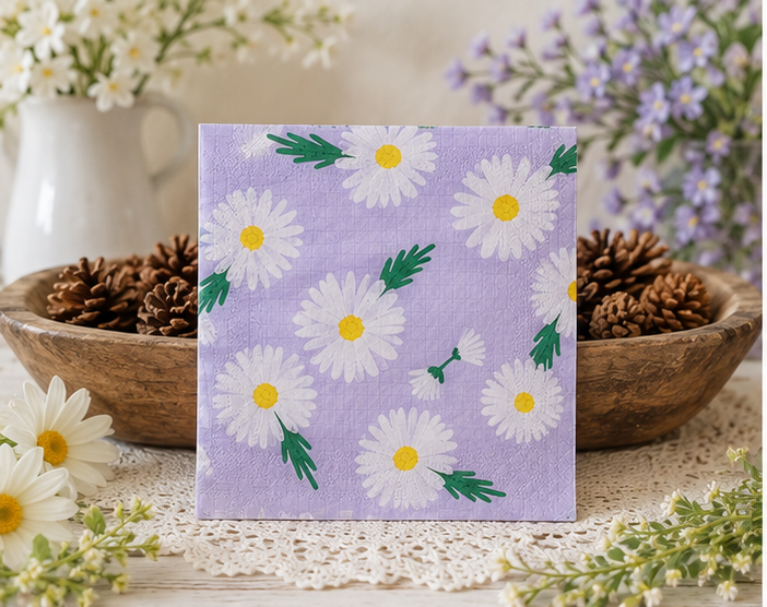 A square paper napkin with a light purple background and a white daisy pattern with green leaves and yellow centers, displayed on a table runner with pinecones and flowers in the background. Great for crafts