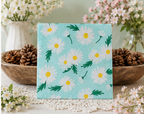 A square napkin print featuring a daisy pattern on a light blue background, styled on a white lace doily with pinecones and white and pink flowers in the background. Great for crafts