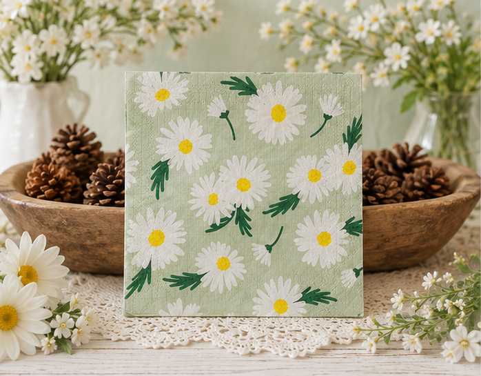 A square paper napkin featuring a daisy pattern, displayed upright on a lace doily with bowls of pinecones and white flowers in the background. Great for crafts