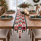 A festive Christmas table runner with a patchwork design of red, green, and cream squares featuring snowflakes, Christmas trees, and reindeer, styled on a wooden dining table with place settings and holiday decorations.