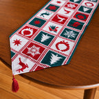 A Christmas-themed table runner with a red, green, and white holiday pattern, featuring a tassel at one end, displayed on a wooden table.