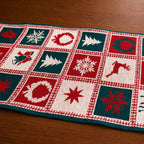 A knitted Christmas-themed table runner laid diagonally on a wooden tabletop. The runner features a grid pattern of red, dark teal, and cream squares with festive motifs including snowflakes, Christmas trees, wreaths, a reindeer, and stars.
