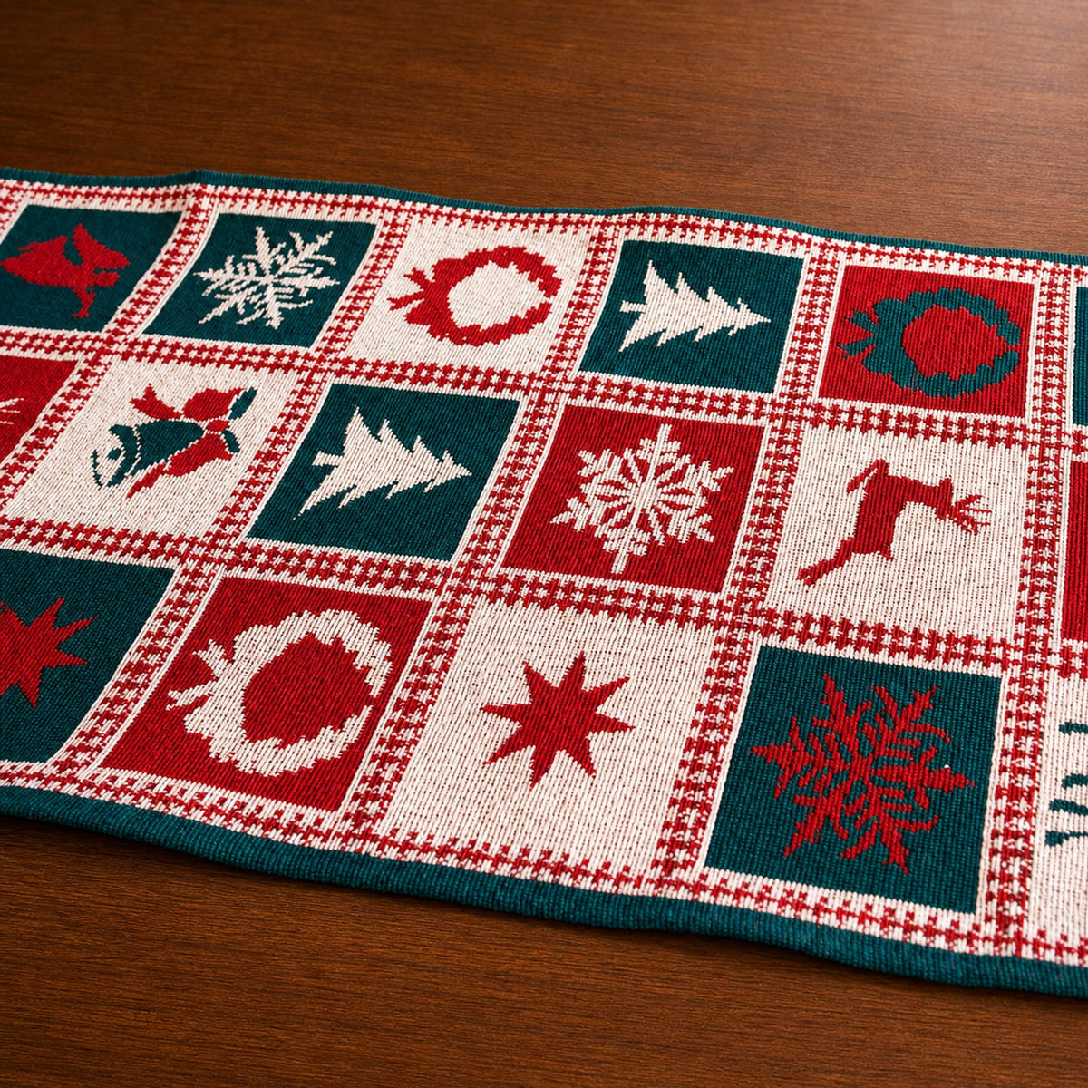 A knitted Christmas-themed table runner laid diagonally on a wooden tabletop. The runner features a grid pattern of red, dark teal, and cream squares with festive motifs including snowflakes, Christmas trees, wreaths, a reindeer, and stars.
