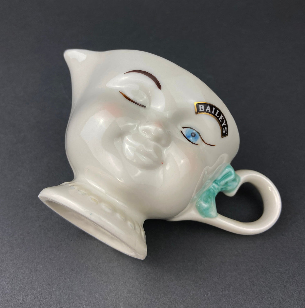 White vintage Bailey’s ceramic creamer shaped like a smiling face with one eye winking, a blue bow, and a small handle.