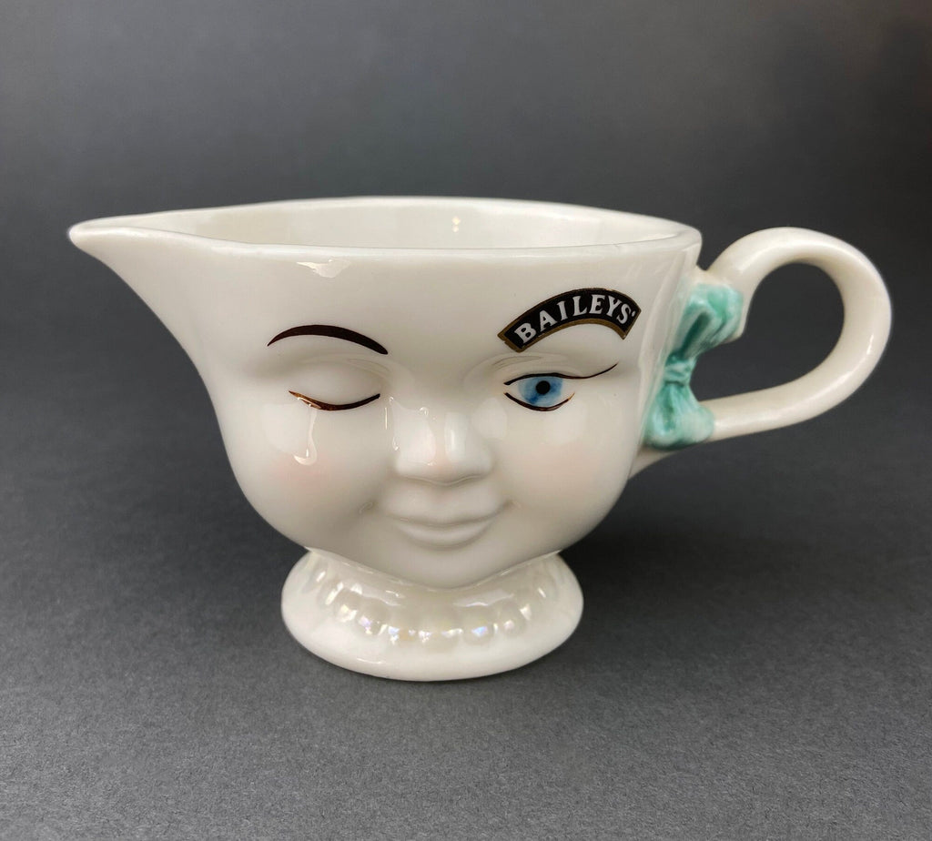 White vintage Bailey’s ceramic creamer shaped like a smiling face with one eye winking, a blue bow, and a small handle.