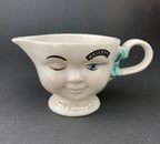 White vintage Bailey’s ceramic creamer shaped like a smiling face with one eye winking, a blue bow, and a small handle.