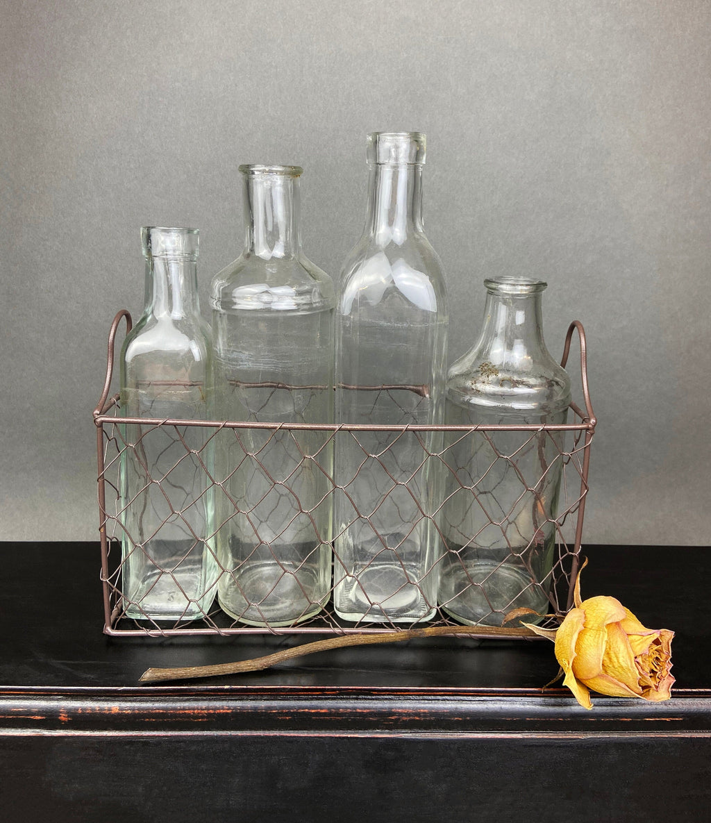 Four vintage clear glass apothecary bottles in a weathered metal basket, styled with yellow flowers.