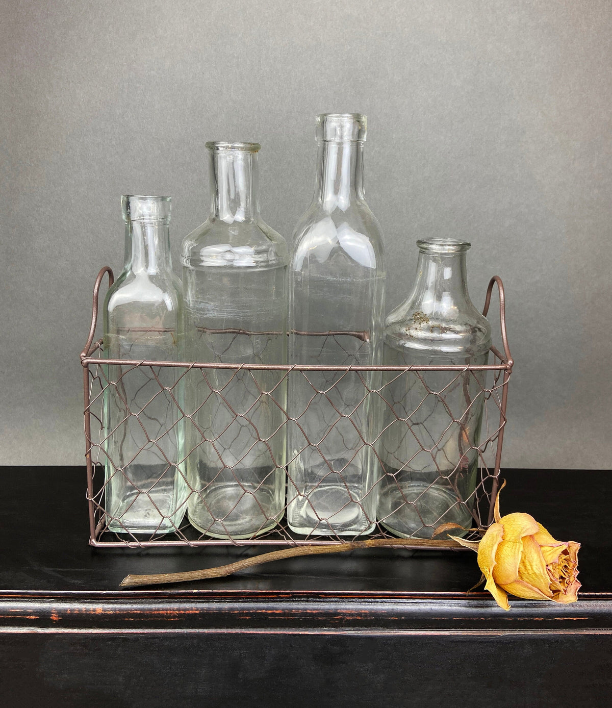 Four vintage clear glass apothecary bottles in a weathered metal basket, styled with yellow flowers.
