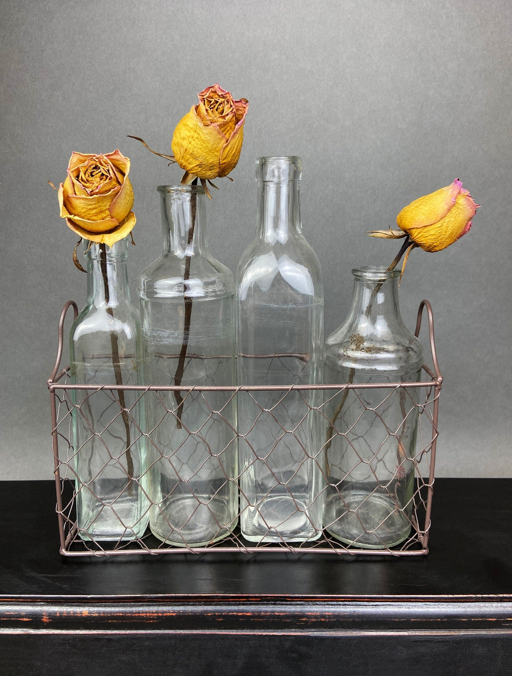 Four vintage clear glass apothecary bottles in a weathered metal basket, styled with yellow flowers.
