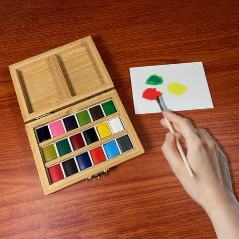 1. Hands holding an open bamboo watercolor box with 18 paint pans and mixing tray
