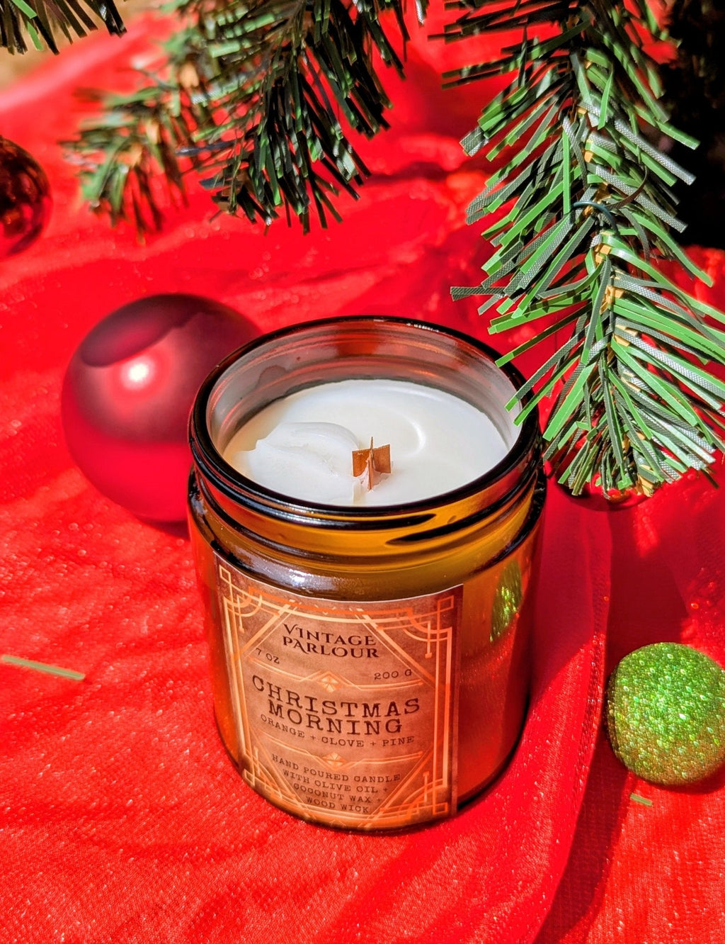 Vintage Parlour Christmas Morning wooden wick candle in an amber glass jar displayed with evergreen branches and colorful Christmas ornaments