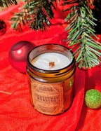 Vintage Parlour Christmas Morning wooden wick candle in an amber glass jar displayed with evergreen branches and colorful Christmas ornaments